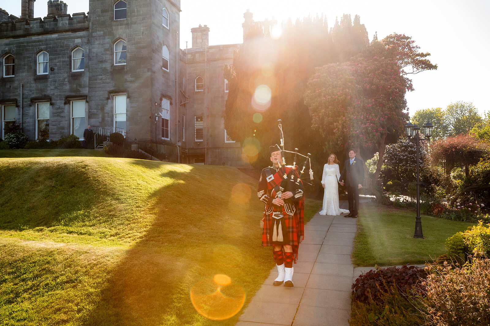 A bagpiper in traditional Scottish attire plays on a garden path, leading a bride and groom near the historic stone walls of Dundas Castle in bright sunlight.